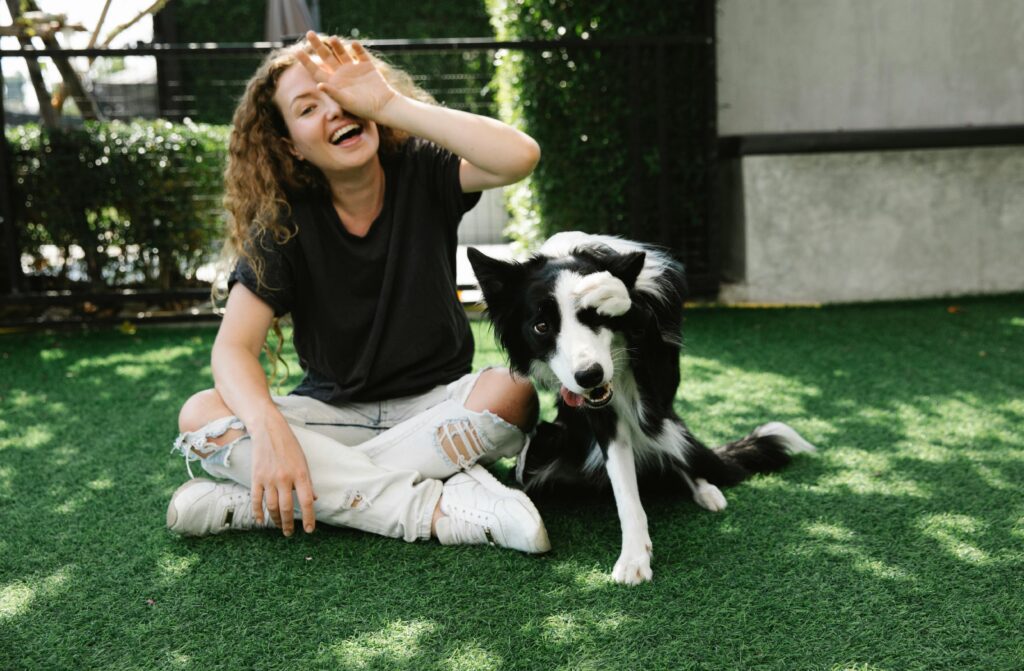 pexels-photo-7210692-7210692 Cheerful female covering eye while sitting with crossed legs against purebred dog and looking at camera on meadow in sunlight
